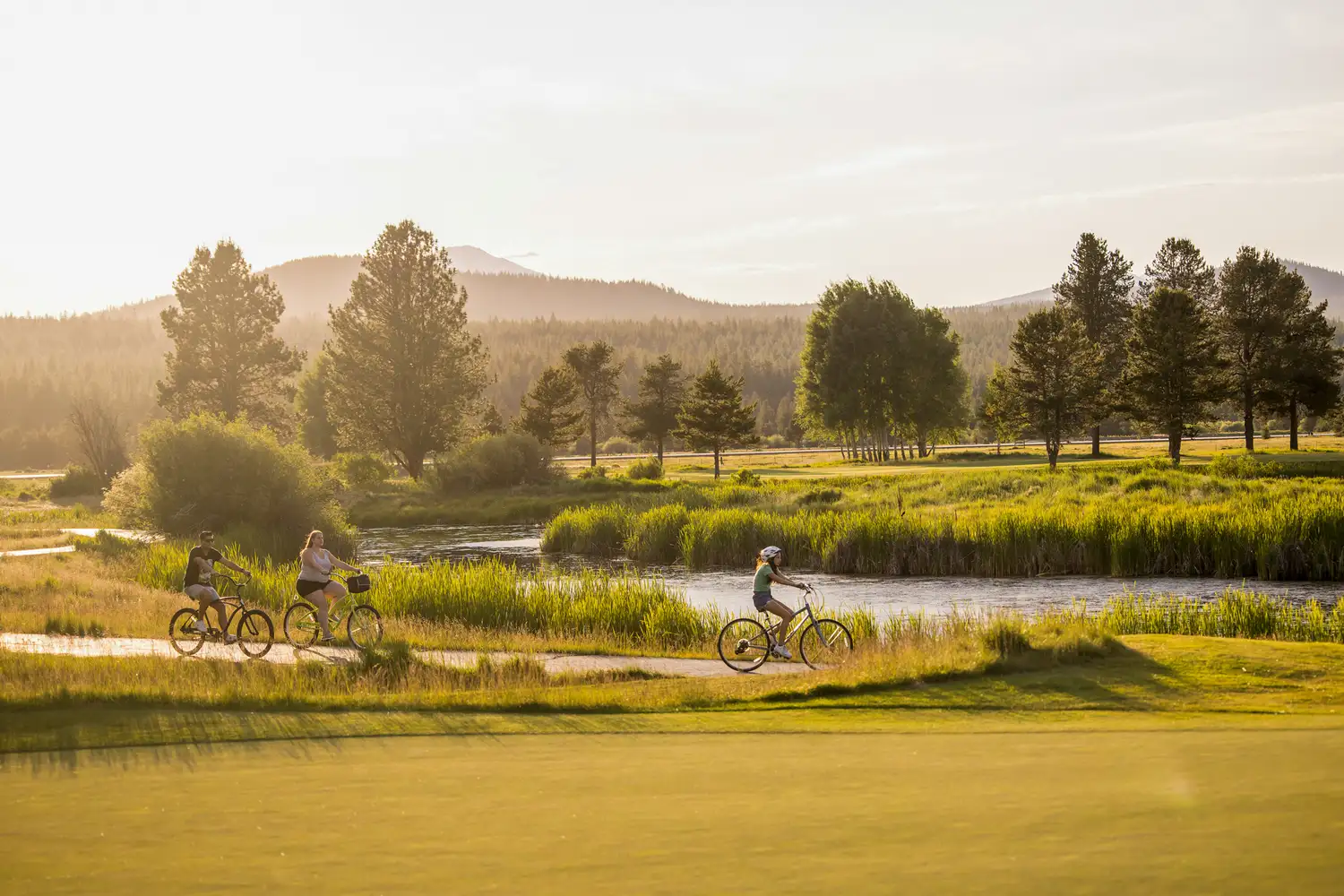 Families riding bicycles along the paved paths of Sunriver Resort next to a winding river.