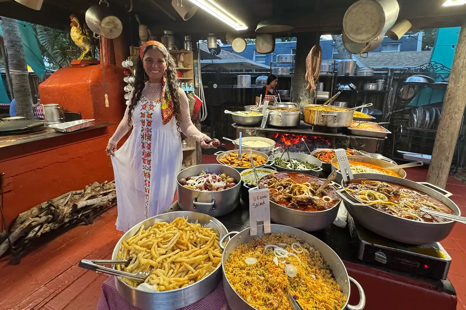 Regina Rodrigues standing behind a table filled with various pans of traditional Brazilian food in an outdoor setting.