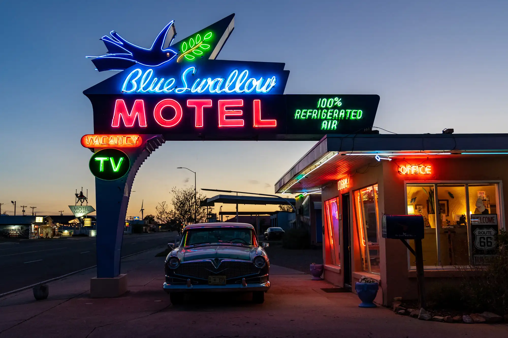Glowing neon sign of the Blue Swallow Motel at dusk with a classic car parked in front.