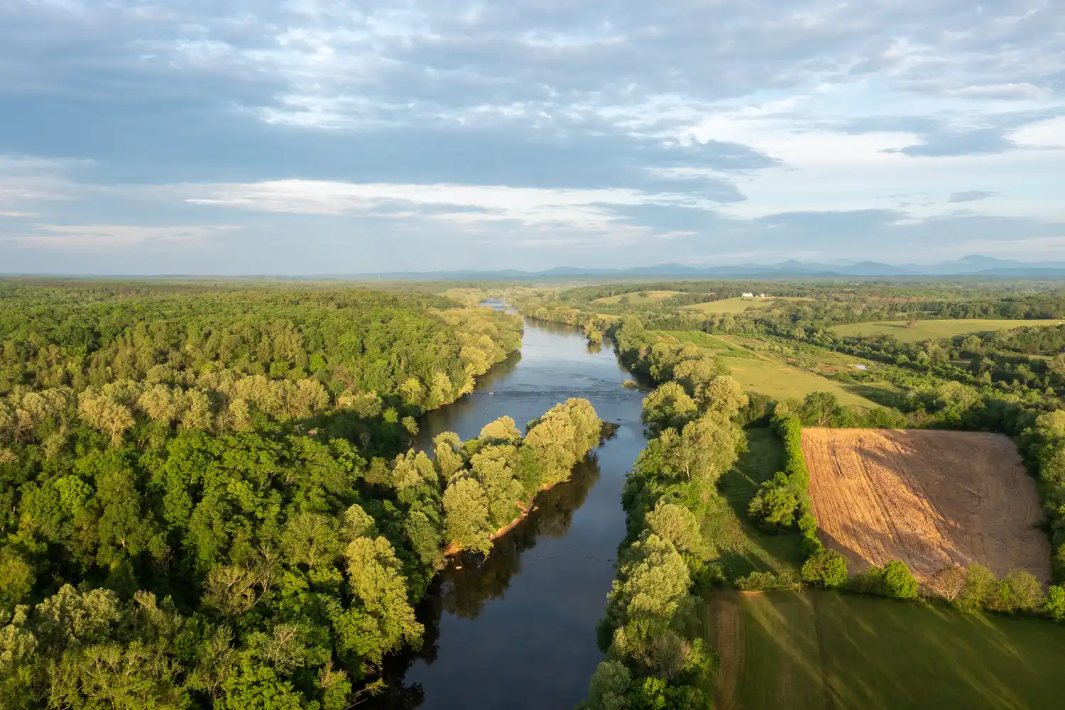Aerial view of a river flowing through a forested and agricultural landscape under a cloudy sky.