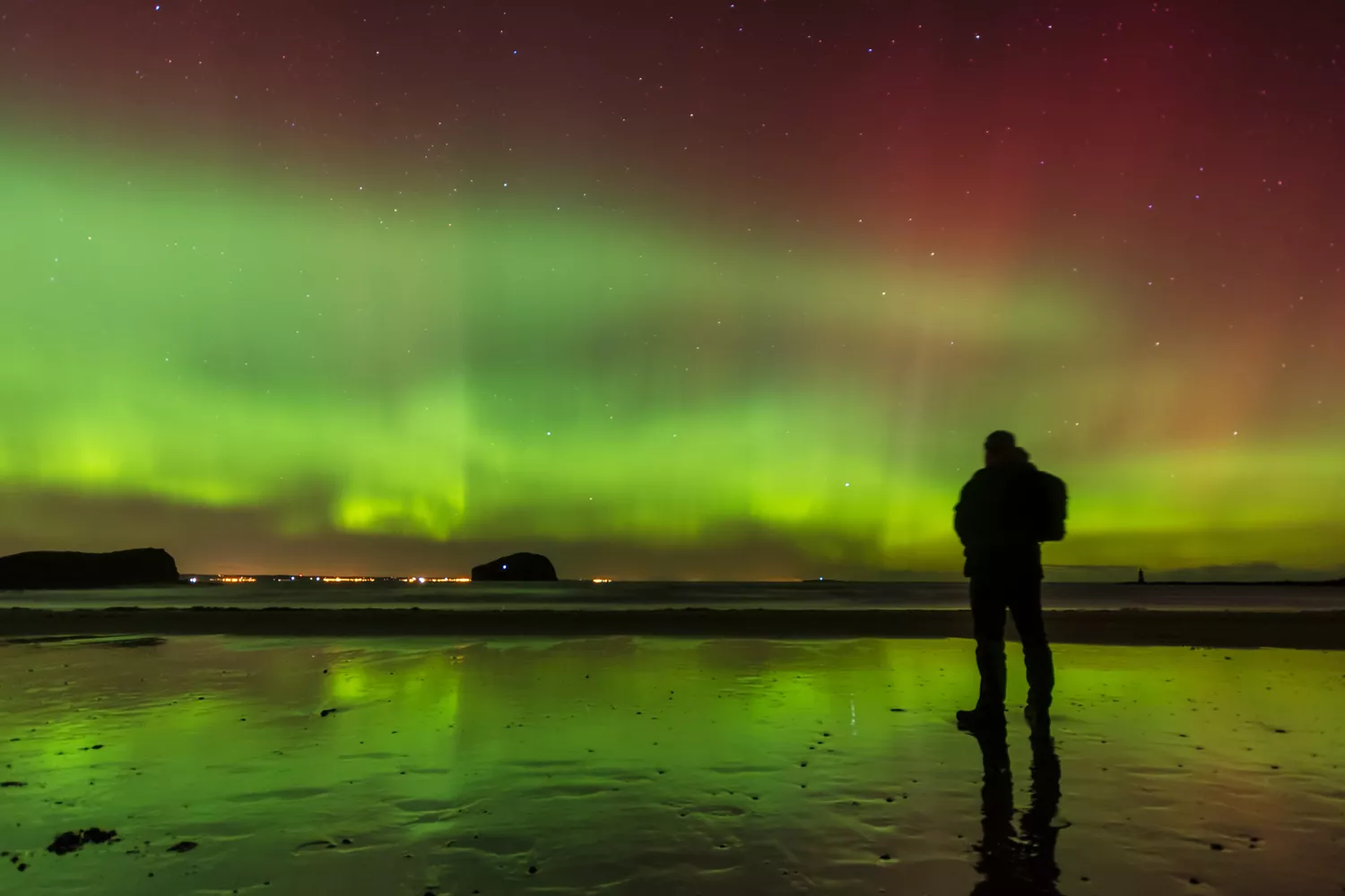 A person silhouetted against the green glow of the Northern Lights on a Scottish beach.