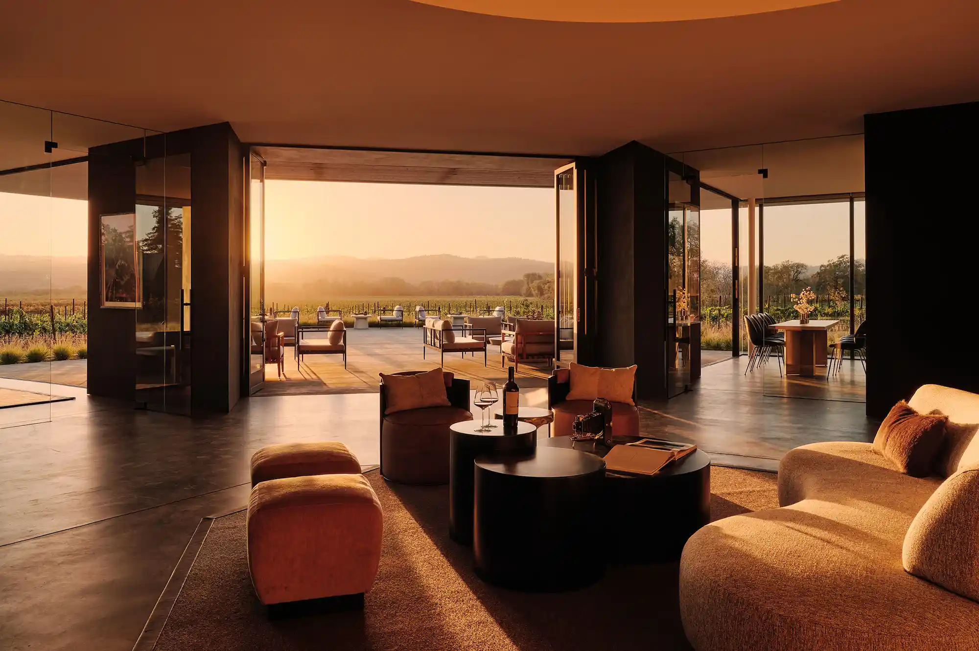 A modern glass-walled tasting room with lounge seating overlooking a sunset vineyard view.