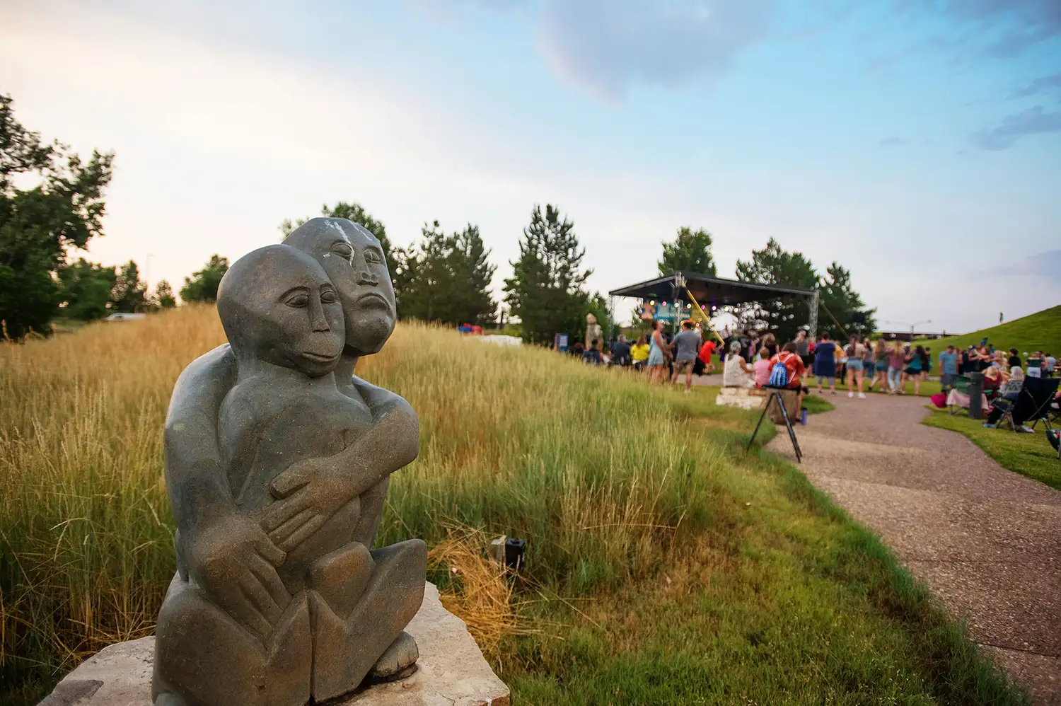 The 'Grandson Protecting Grandfather' stone sculpture in a park setting with visitors and a pavilion in the background.