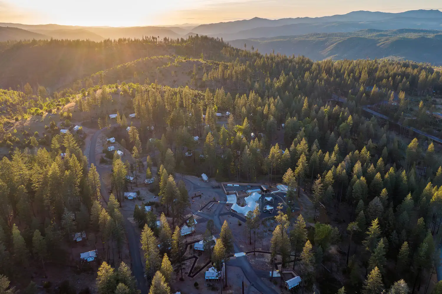 Aerial drone view of white canvas tents nestled in a thick green forest under a sunset sky.