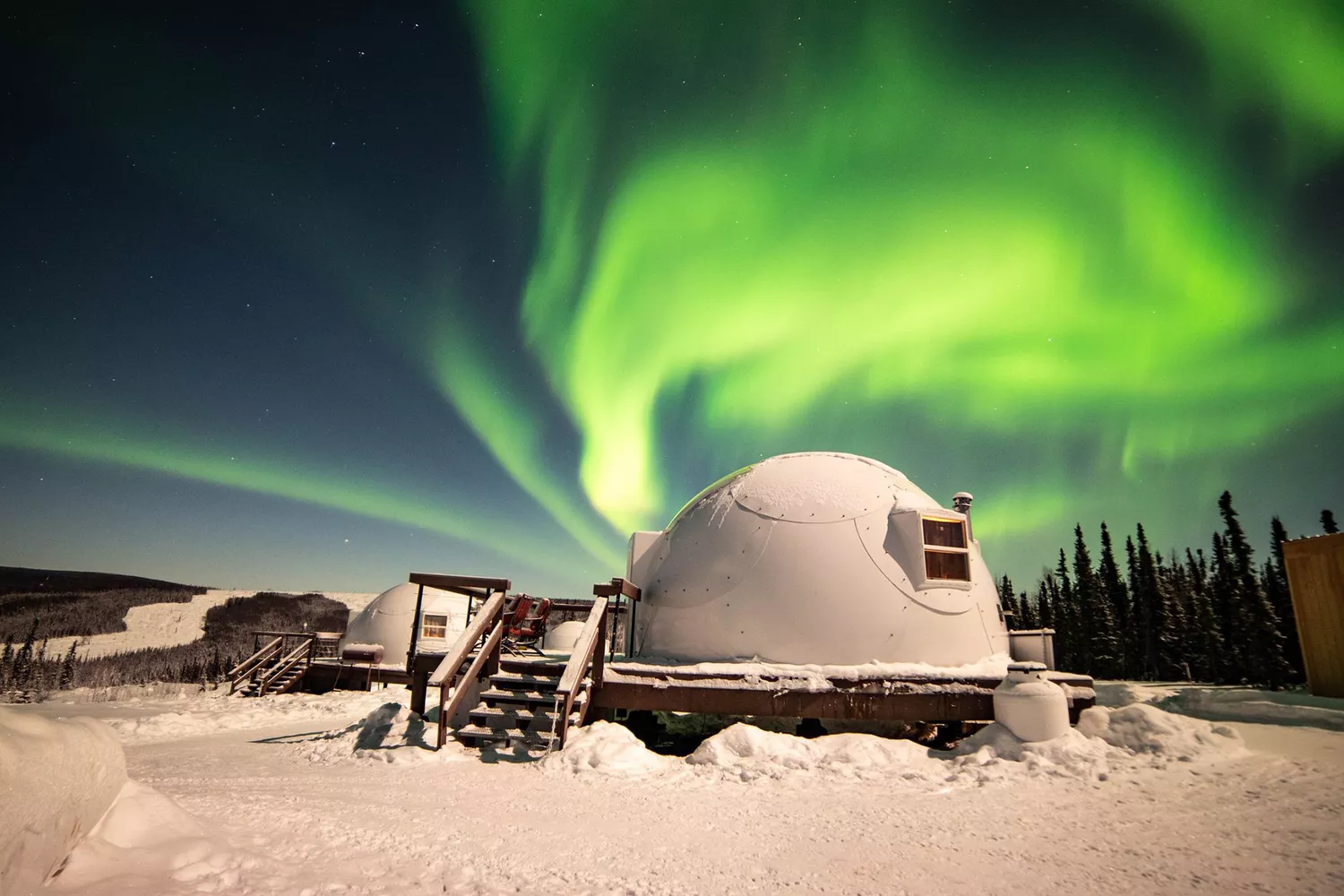 Bright green aurora borealis lights up the night sky above a glowing geodesic dome in Alaska.