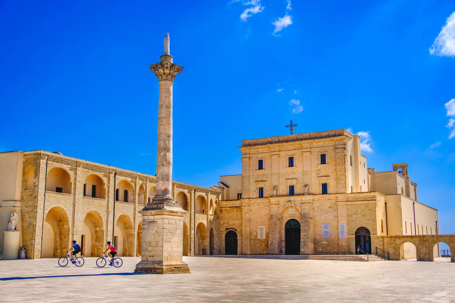 Cyclists riding through a historic plaza with a column and old buildings in Lecce.