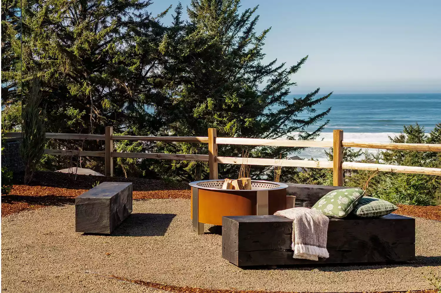 A communal fire pit area with wooden benches, pillows, and blankets under the forest canopy.