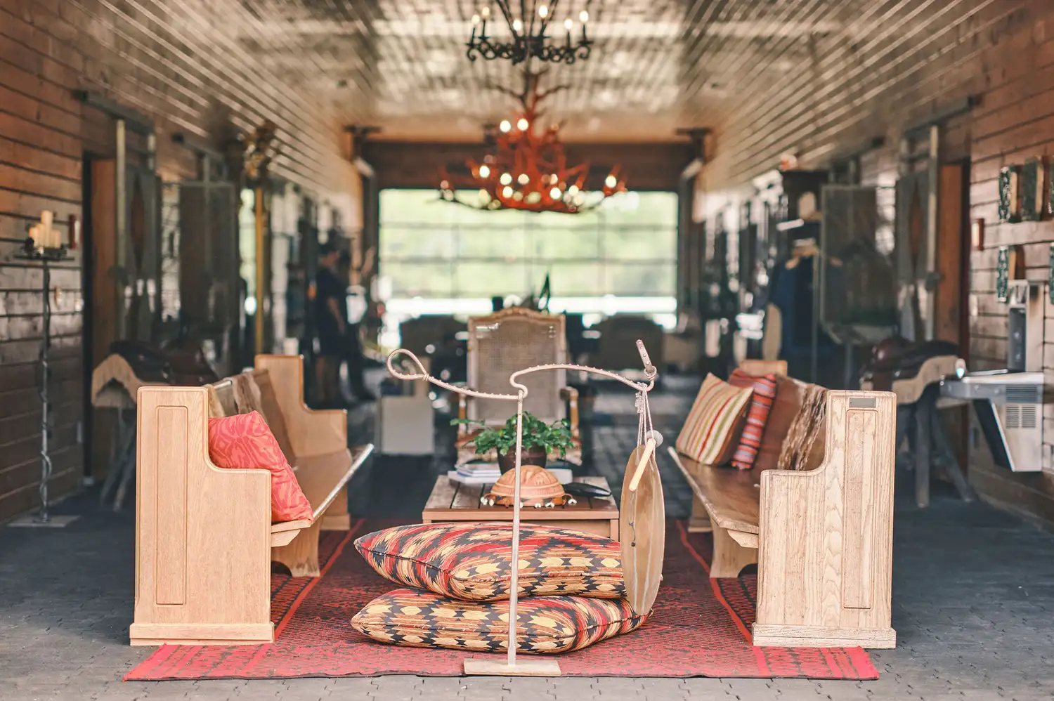 A rustic lounge area inside a converted stable featuring wooden benches, a central table, and a chandelier.