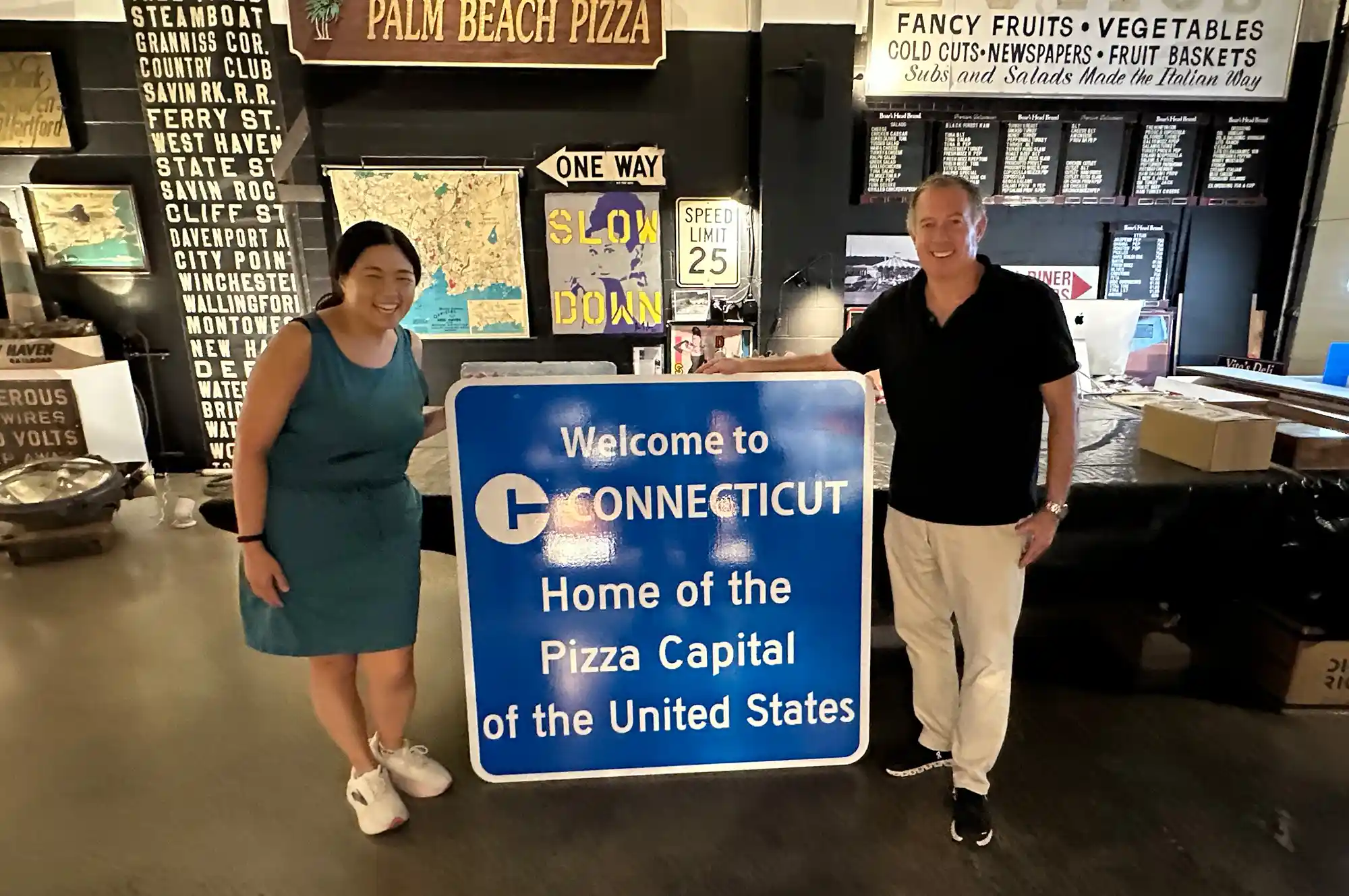 Two people standing next to a sign that reads 'Welcome to Connecticut Home of the Pizza Capital of the United States'.