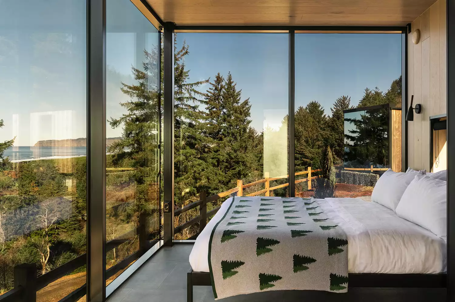 Interior view of a mirrored cabin showing modern furniture and a panoramic view of the forest through floor-to-ceiling glass.