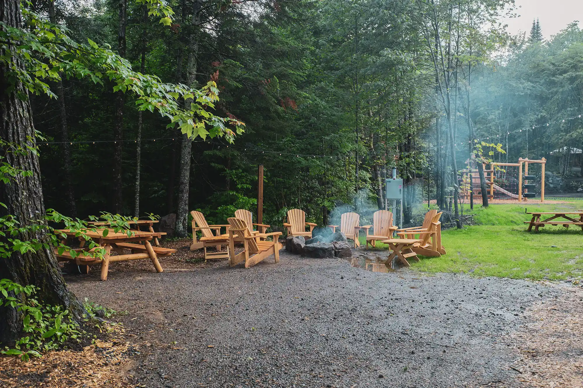 A circular outdoor fire pit with wooden chairs surrounded by lush trees and a playground in the distance.