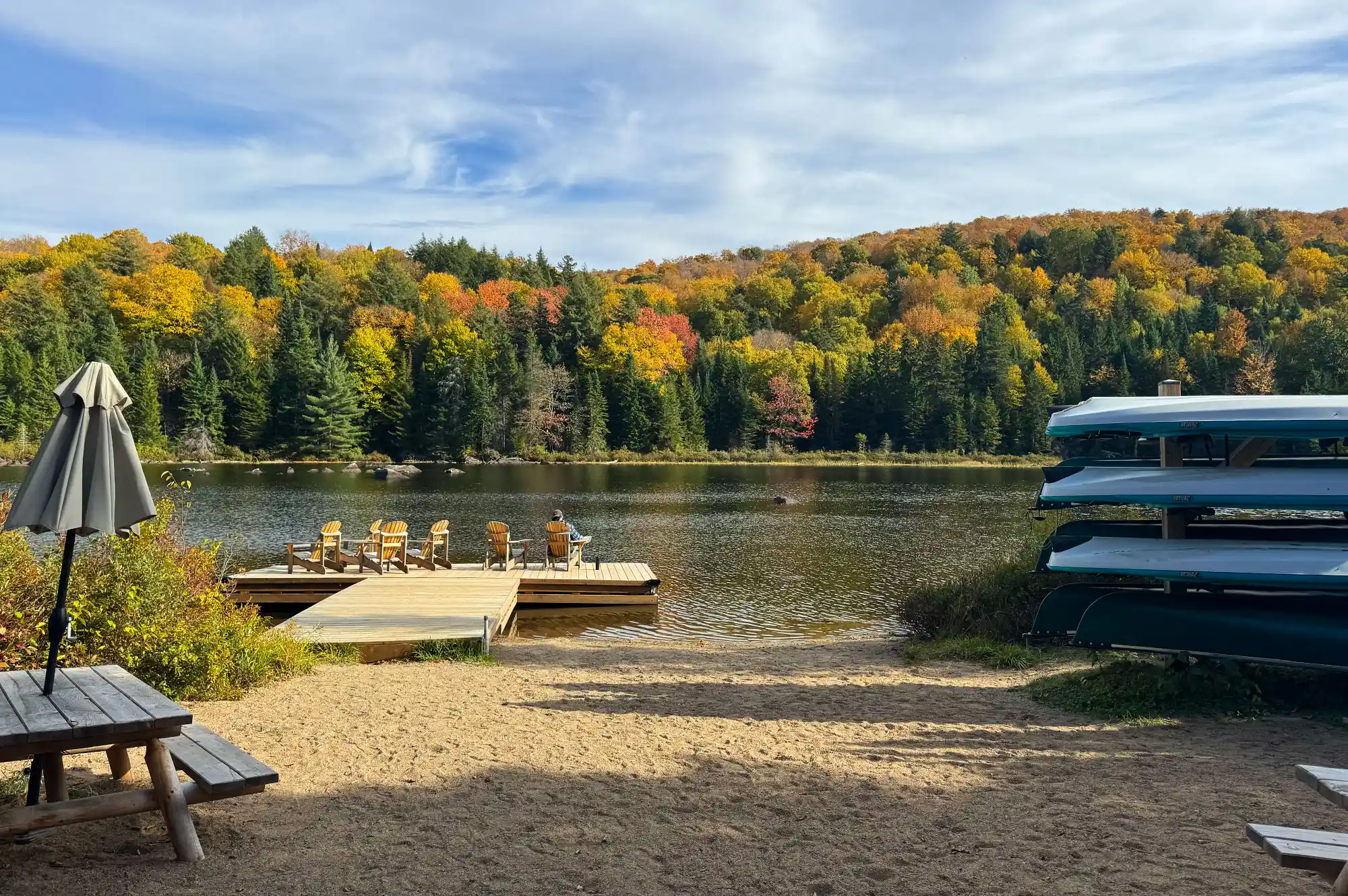 A sandy beach at Lac à la Truite with stacked canoes, a wooden dock, and picnic tables under autumn foliage.