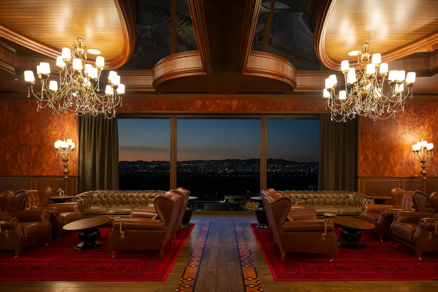 A luxury lounge with dark leather chairs and chandeliers overlooking a city view at night.