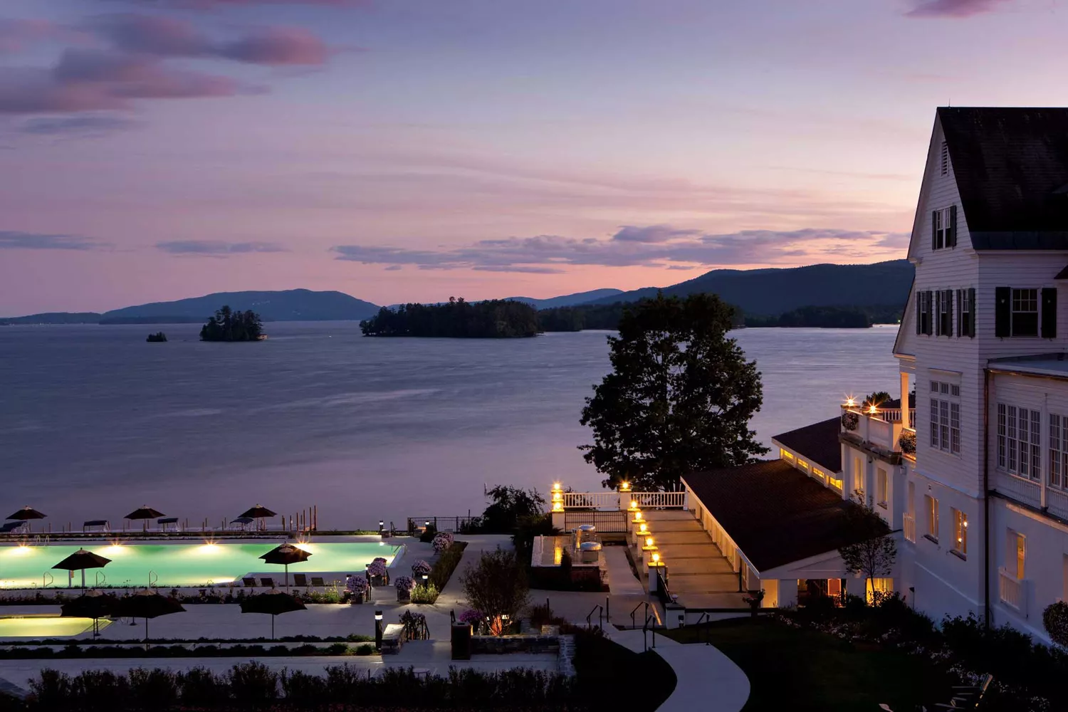 Sunset view of The Sagamore Resort on Lake George featuring the swimming pool and illuminated guest buildings.