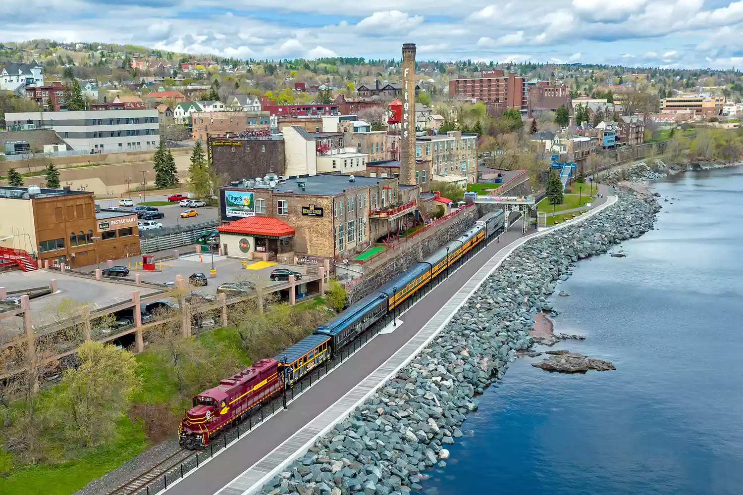 Aerial view of the Duluth Zephyr train traveling along the rocky shoreline of Lake Superior near wooded neighborhoods.
