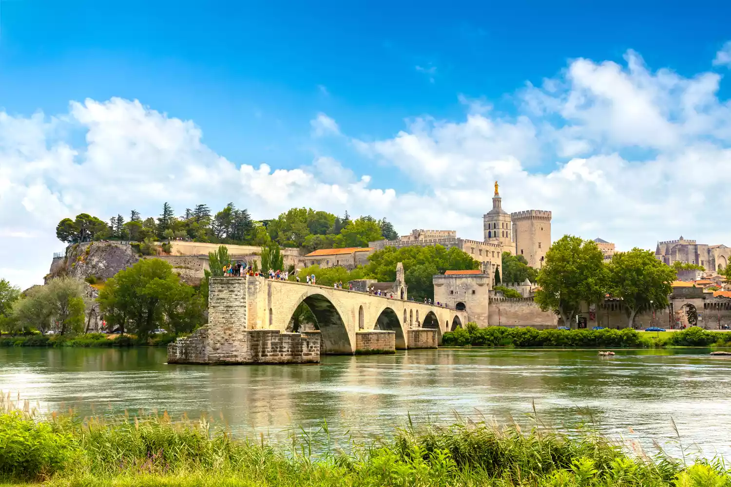 The remaining arches of the Saint Benezet bridge extending into the Rhone river under a blue sky.