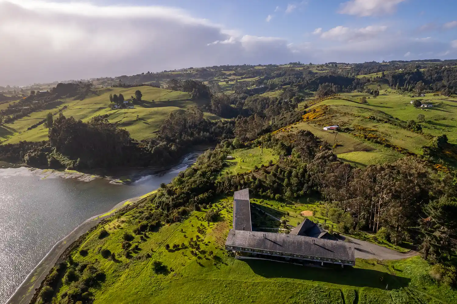 Aerial view of a contemporary wooden hotel building situated on a grassy hill overlooking Pullao Bay in Chiloé.