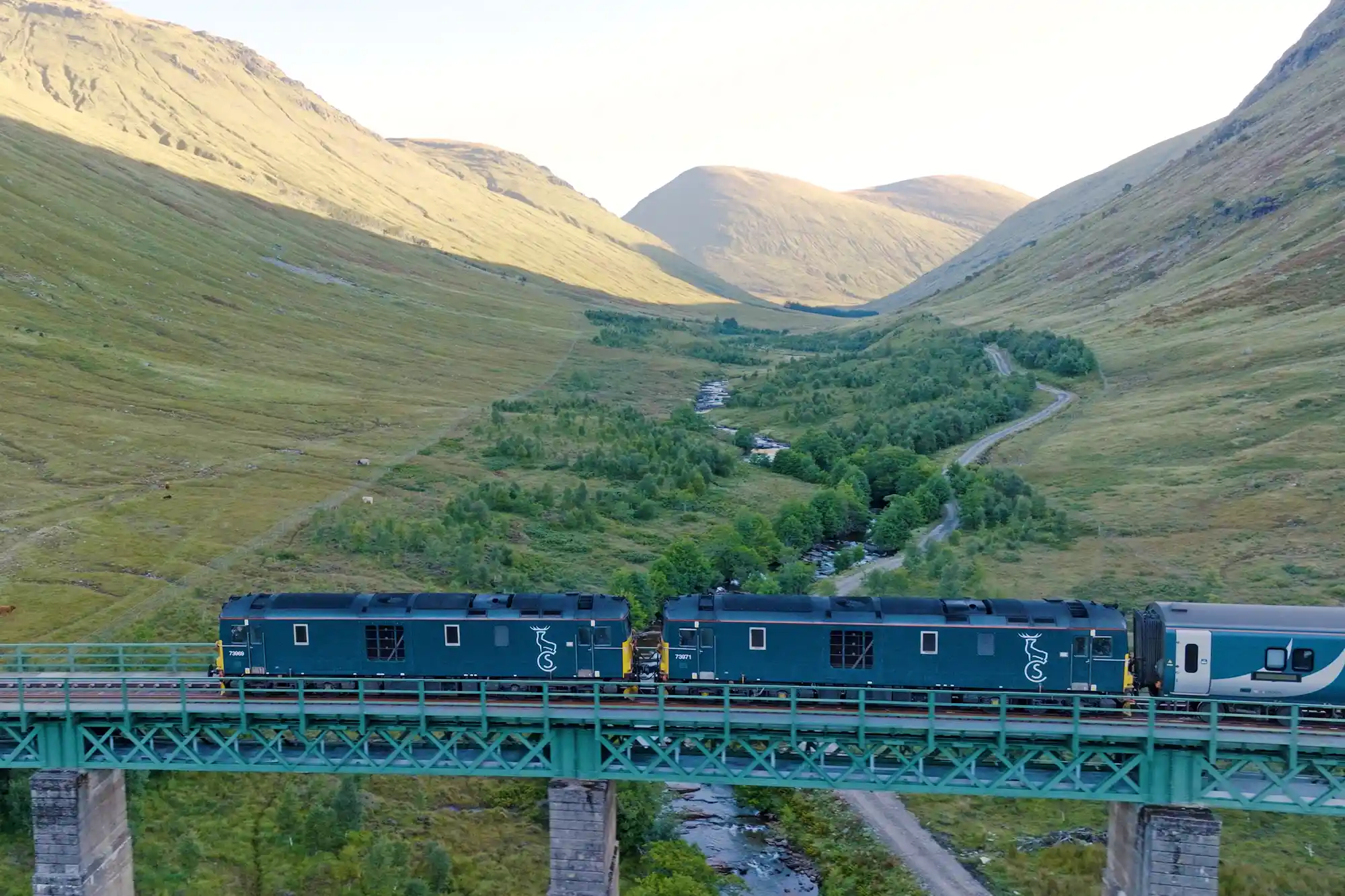 The Caledonian Sleeper train crossing a stone viaduct surrounded by green hills and misty mountains.