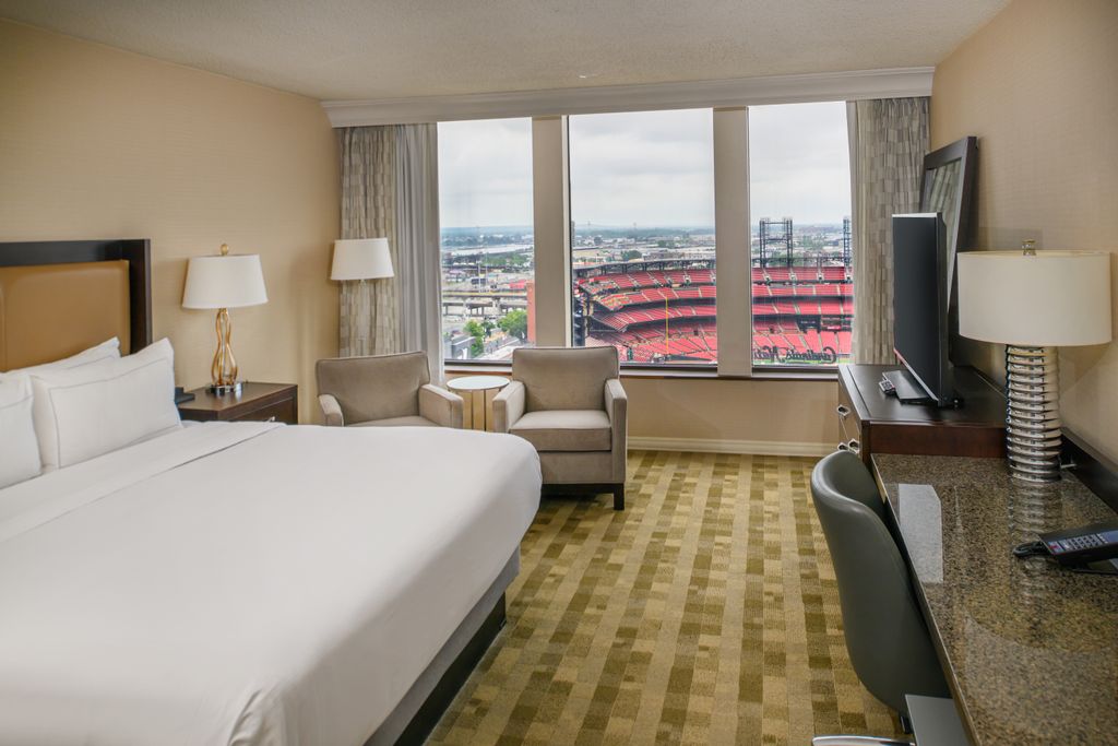 A hotel room with a bed positioned to face a large window overlooking Busch Stadium.