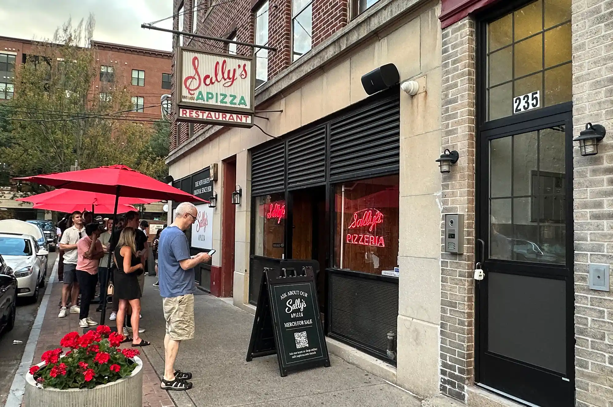 A long queue of people waiting outside the brick storefront of Sally's Apizza.