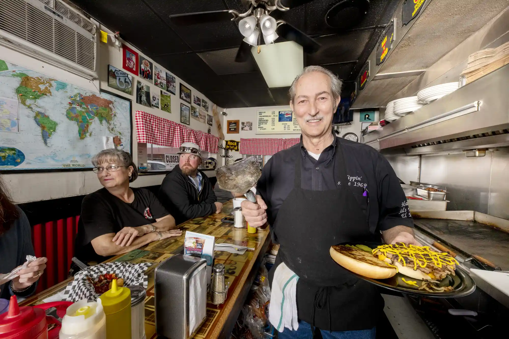 A restaurant owner behind a wooden diner counter serving a burger to patrons.