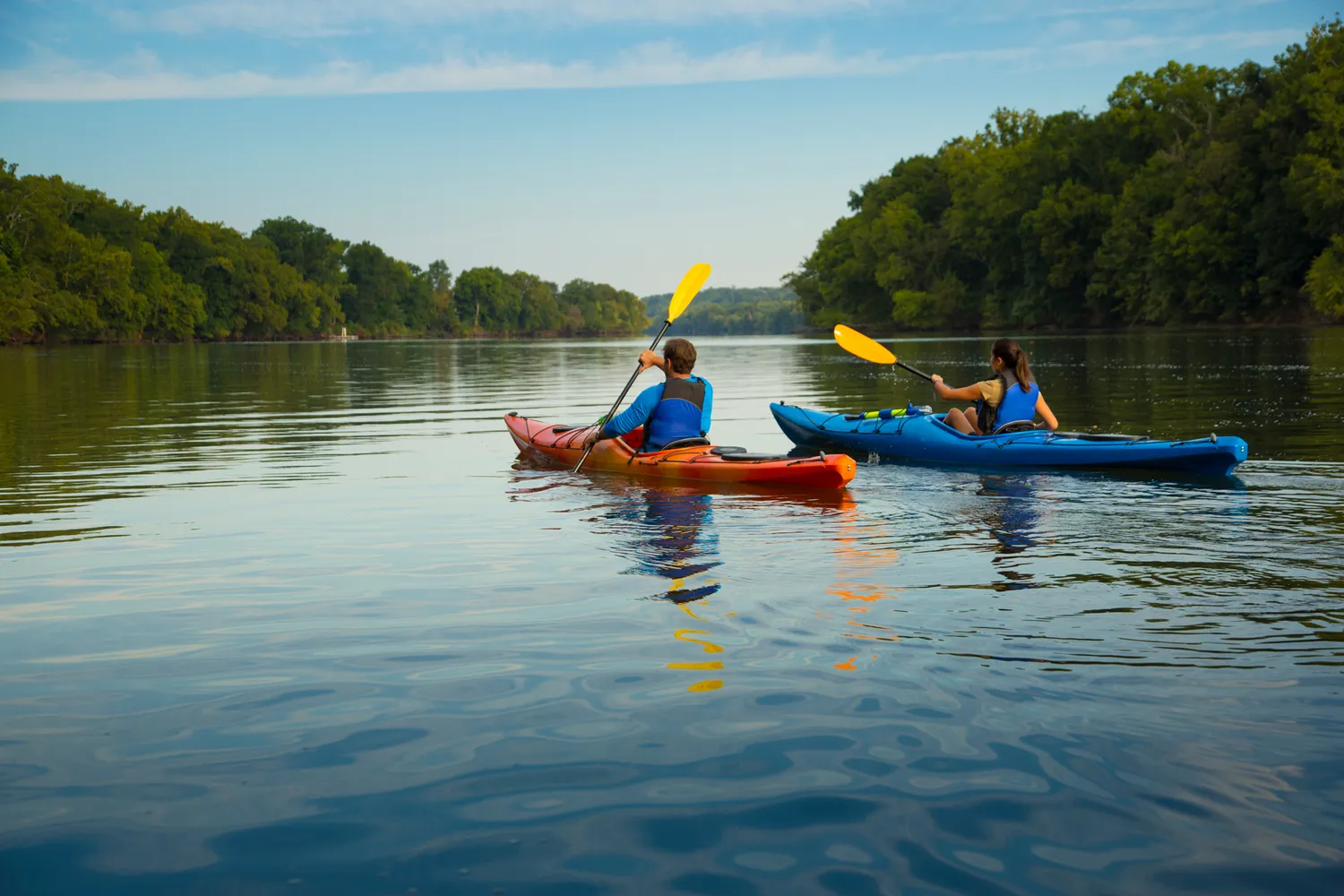 Two people kayaking on a calm river surrounded by trees.