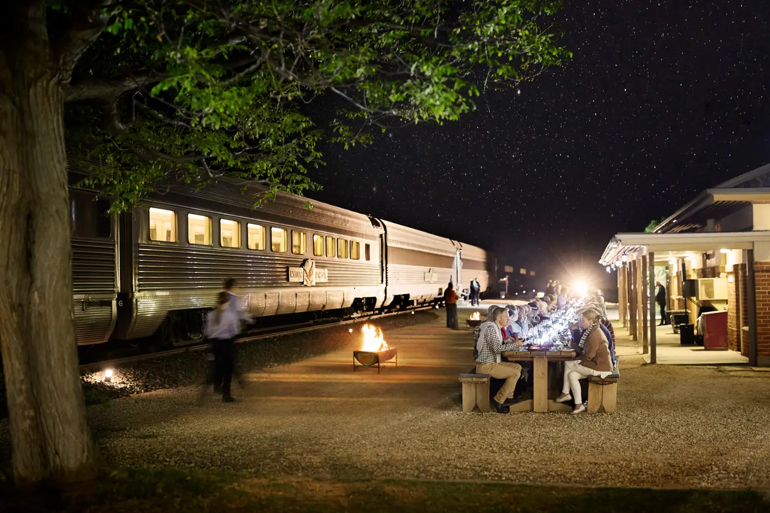 A group of people dining outdoors by a fire pit next to a stopped train at night.
