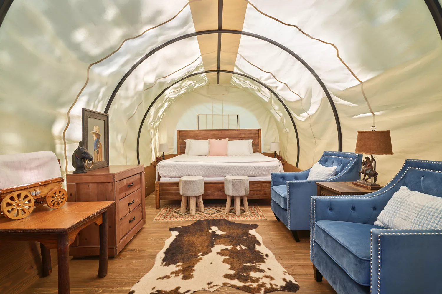 Interior view of a covered wagon showing a wooden bed frame, Western-style textiles, and a chandelier.