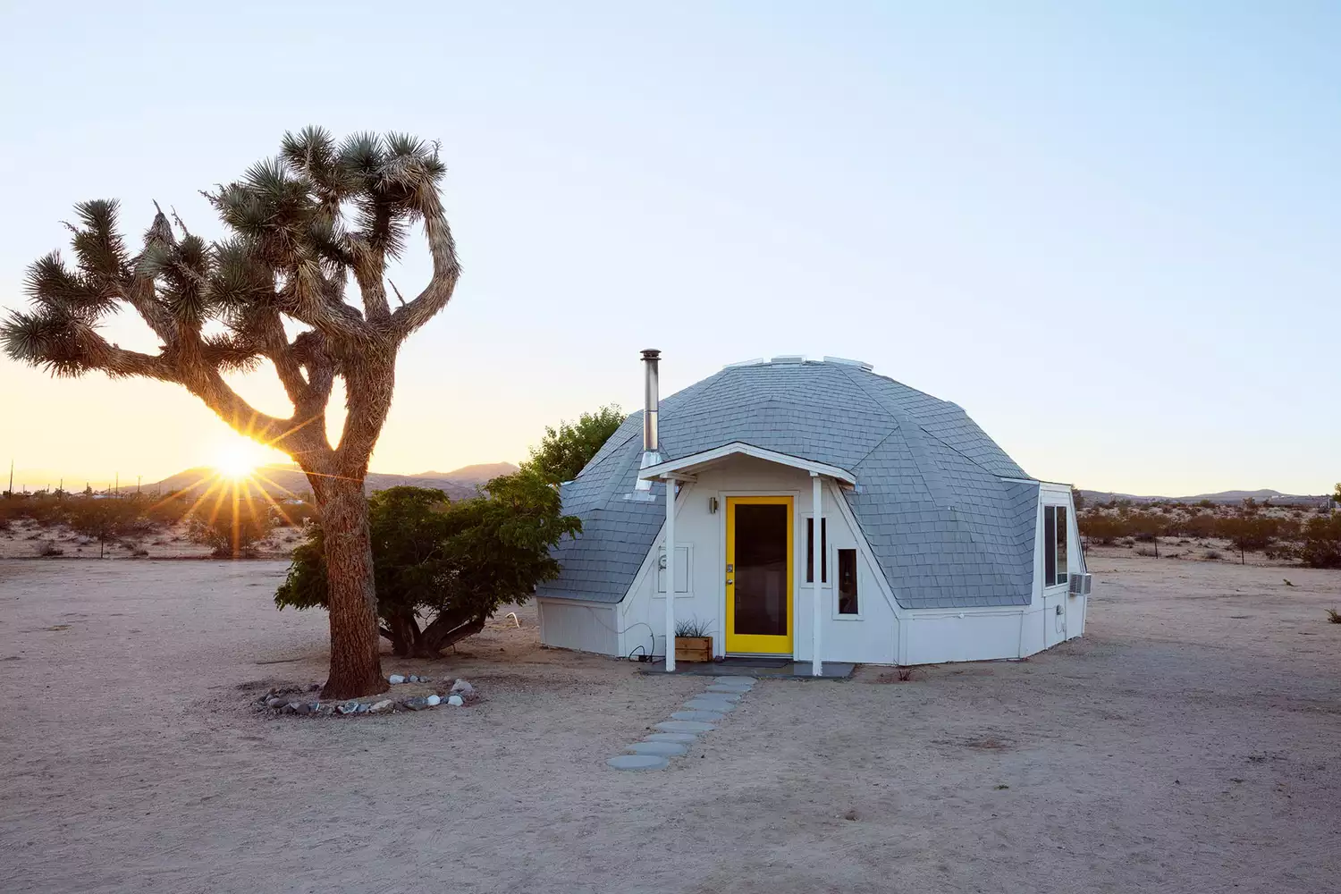Exterior view of a white geodesic dome nestled among desert rocks and scrub in California.