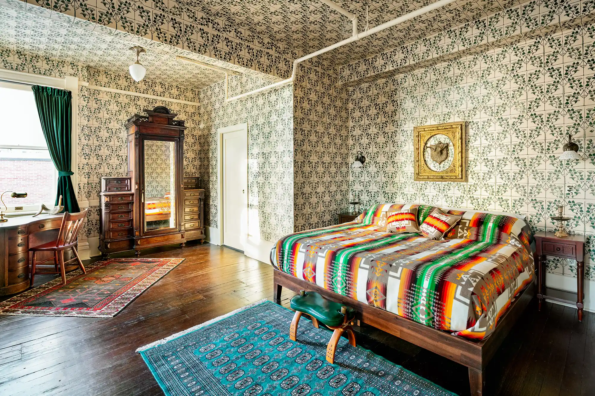 Stylish bedroom with patterned wallpaper, a wooden bed featuring vibrant bedding, and antique furniture accents.