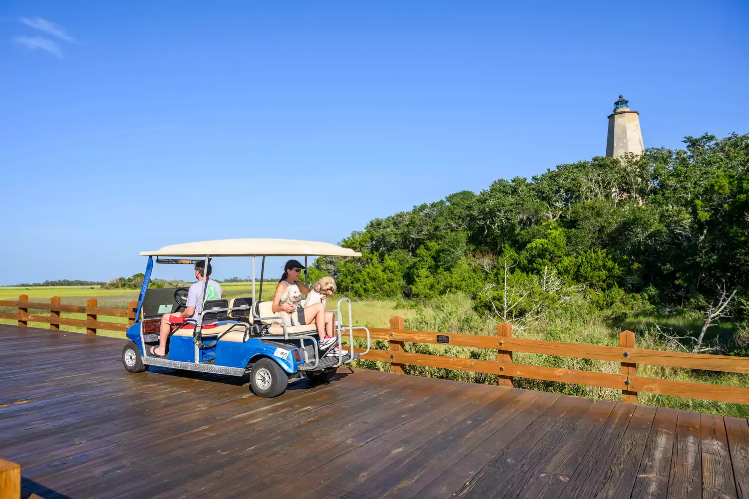 A family on a golf cart crossing a bridge with the Old Baldy lighthouse in the background.