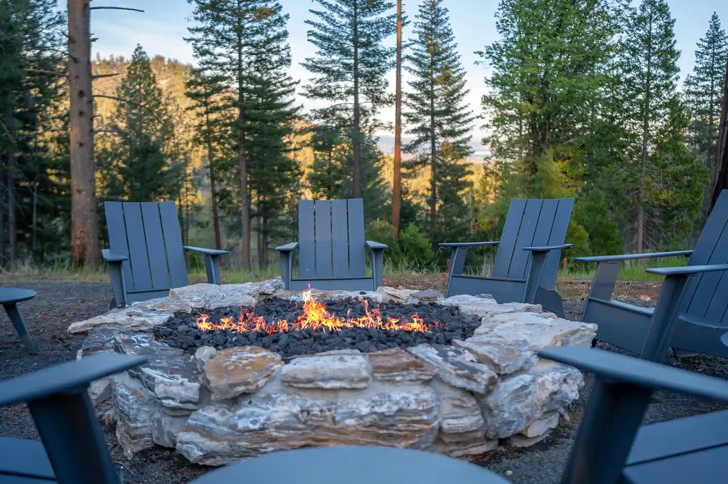 A circular stone fire pit with black Adirondack chairs arranged around it in a wooded clearing.