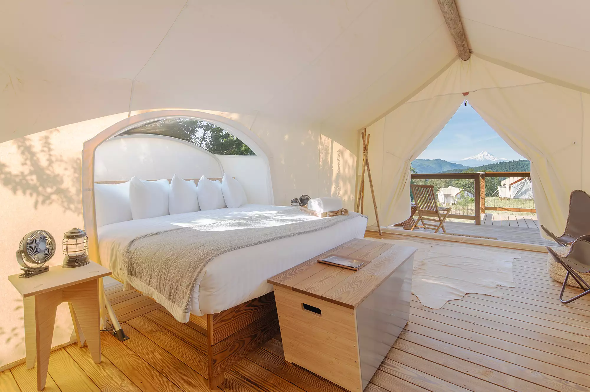 Interior of a canvas tent showing a large bed with white linens and a view through the flap to a private outdoor deck.