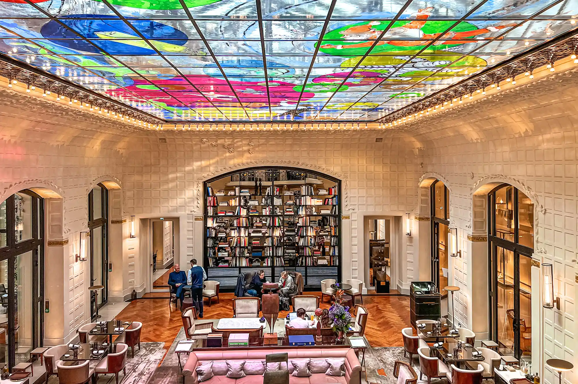 The hotel atrium featuring a colorful glass ceiling and book-filled archway with guests in a lounge area.