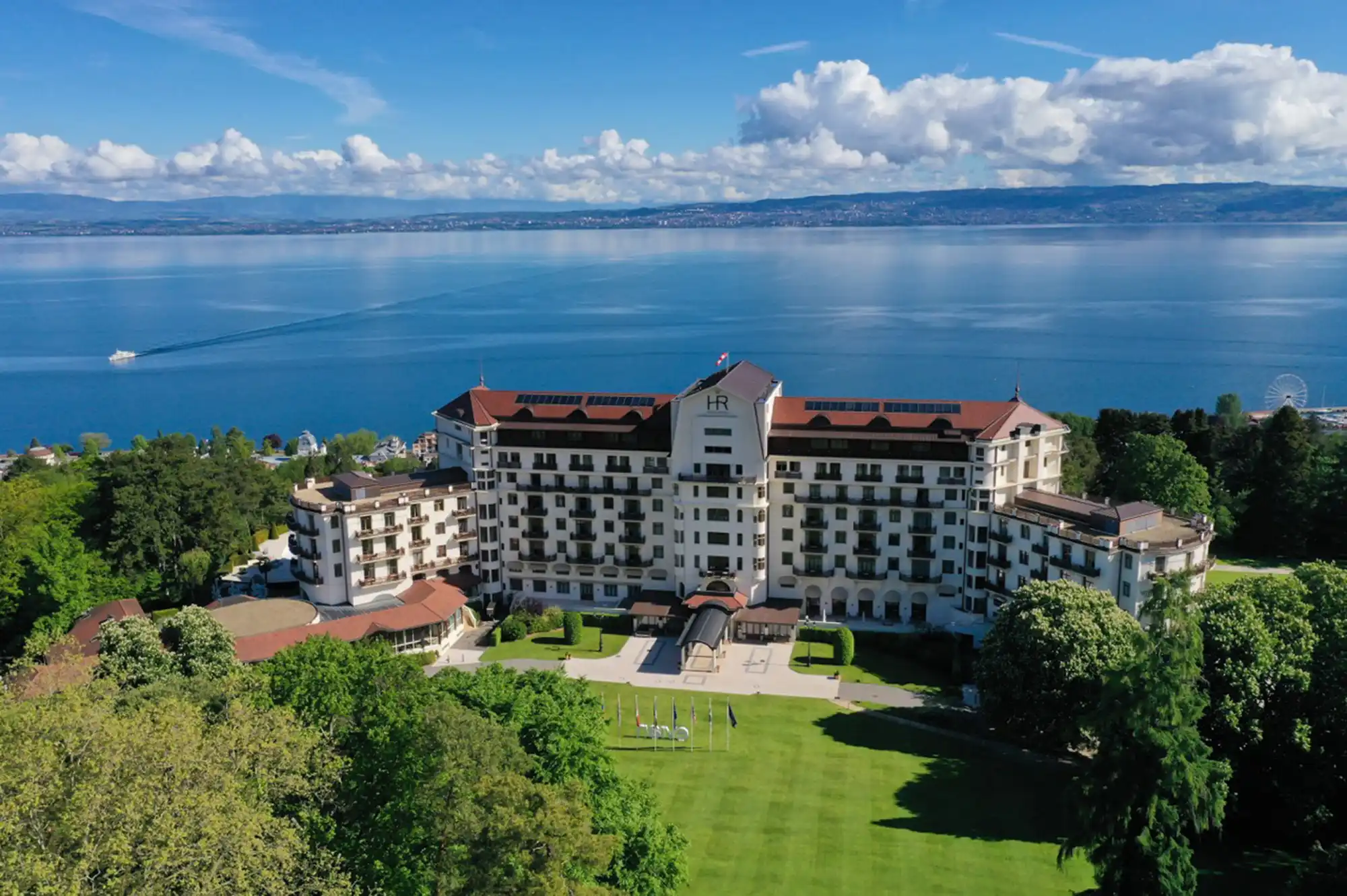 A large, historic luxury hotel building situated on a hillside overlooking a lake and mountains.