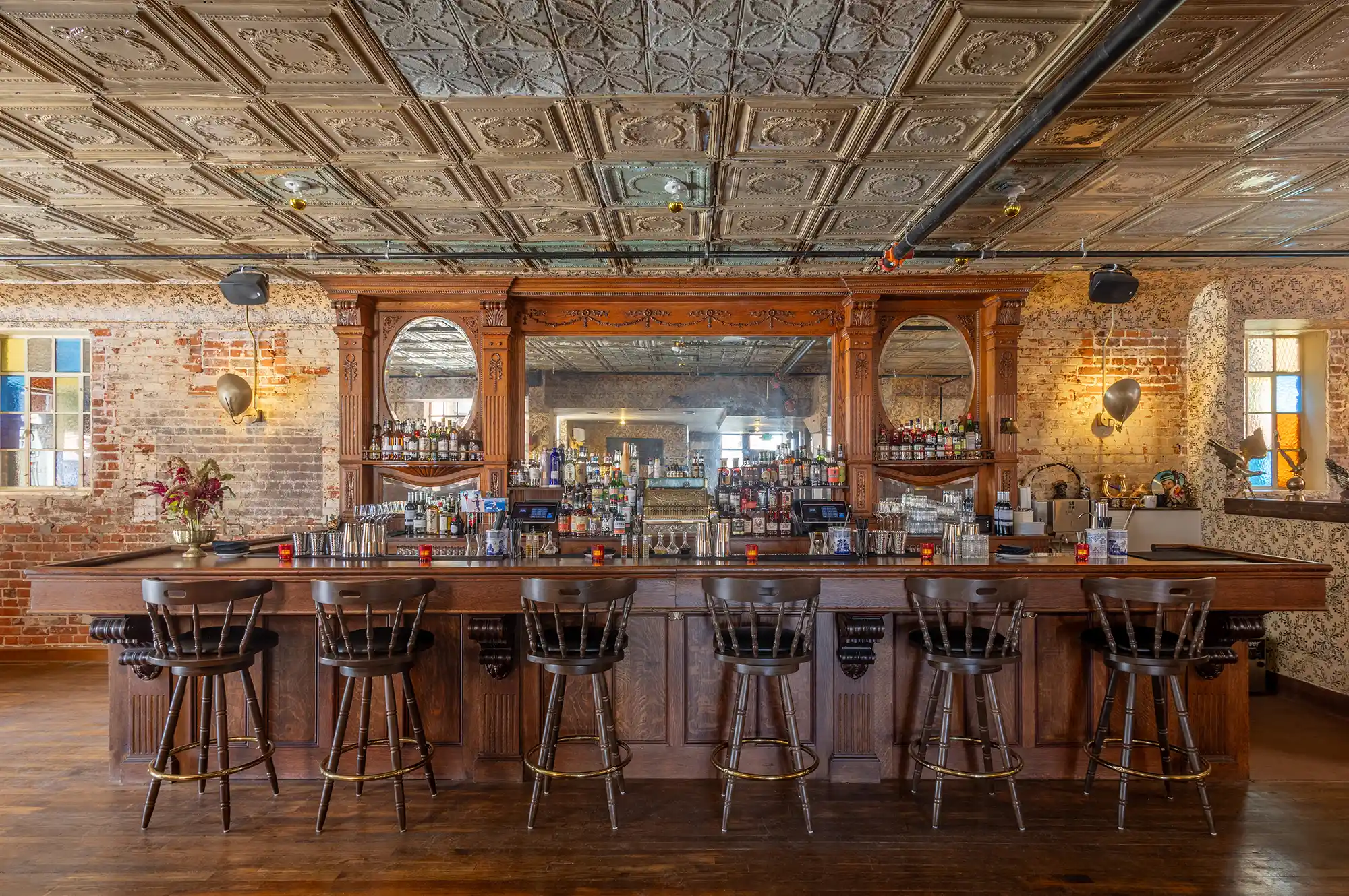 A rustic wooden bar counter with vintage mirrors and curated spirit bottles in a dimly lit setting.